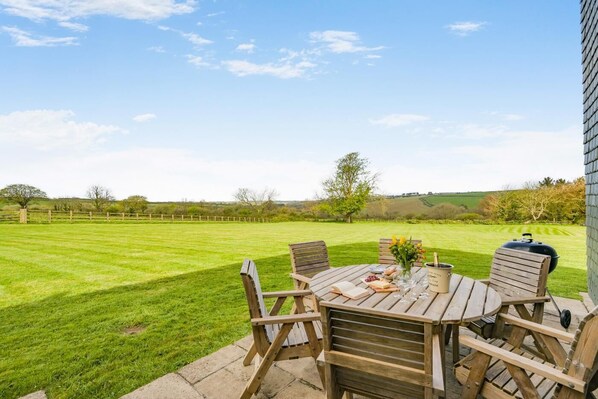 A patio with a table set for dining, a grill, and a view of a grassy field. - 3 Bed in Wadebridge (oc-m34228) (Saint Issey)