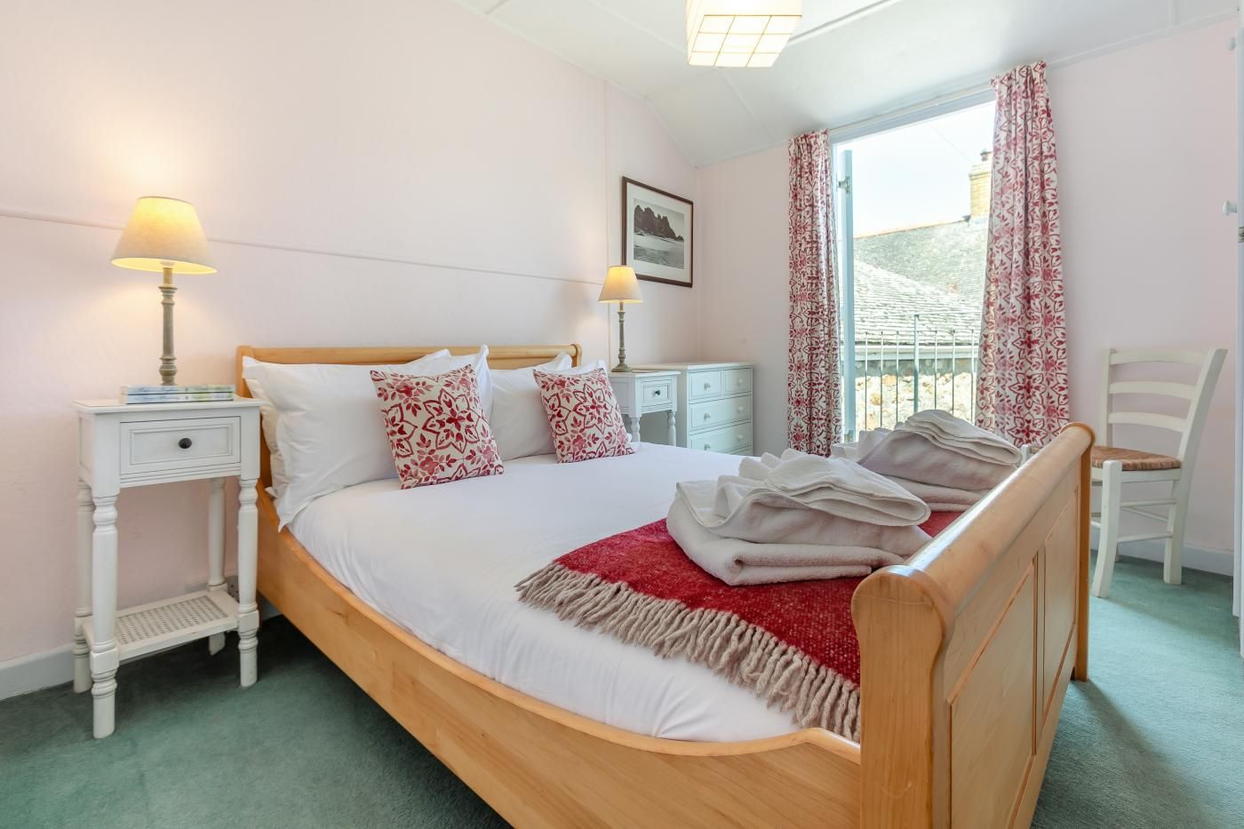 A neatly made bed with a red throw blanket, two bedside lamps, a white nightstand, a white chair, and a window with patterned curtains.