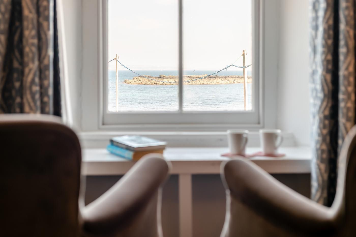 Une pièce avec une fenêtre donnant sur une plage, une chaise, une table avec un livre et deux tasses.
