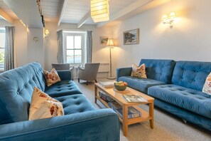A living room with two blue sofas, a coffee table with books and a bowl of fruit, a floor lamp, and a window with curtains. - 5 Bed in Mousehole (oc-kellan) (Mousehole)