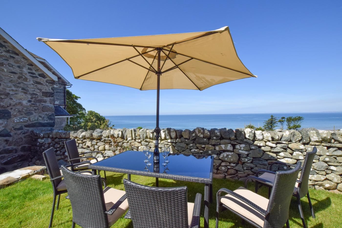 Une table avec quatre chaises et un grand parasol, placée devant un mur de pierre, avec vue sur la mer.