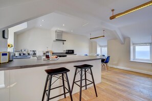 A modern kitchen with a central island, bar stools, and a dining area with a view. - 2 Bed in Barmouth (oc-bow52) (Llanaber)
