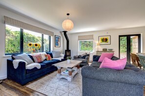 A cozy living room with a blue sofa, a grey armchair, and a coffee table. There is a large window with a view of greenery outside. - 4 Bed in Trevone (oc-p00696) (Trevone)