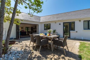 A patio with a table set for breakfast, surrounded by wicker chairs, and a garden area with trees and a white building in the background. - 4 Bed in Trevone (oc-p00696) (Trevone)
