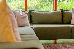 A green sofa with cushions, a window with a view of greenery, and a wooden floor.