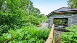 A stone building with a large window, surrounded by lush greenery and a wooden deck. - 2 Bed in Tresarrett (oc-b34255) (Blisland)