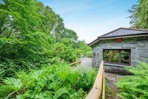 A stone building with a large window, surrounded by lush greenery and a wooden deck.