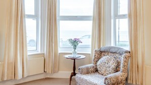 Ein Eckzimmer mit großem Fenster, einem blumigen Sessel und einem kleinen Tisch mit Blumenvase.