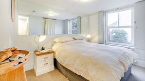 Ein Schlafzimmer mit einem Bett, Nachttischlampen, einem Holznachttisch, einem Spiegel und einem Fenster.