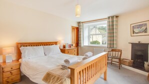 A neatly made bed with a wooden headboard, a bedside lamp, a wooden nightstand, a chair, a fireplace, and a window with curtains. - 2 Bed in Gunnerside (oc-ds1068) (Gunnerside)