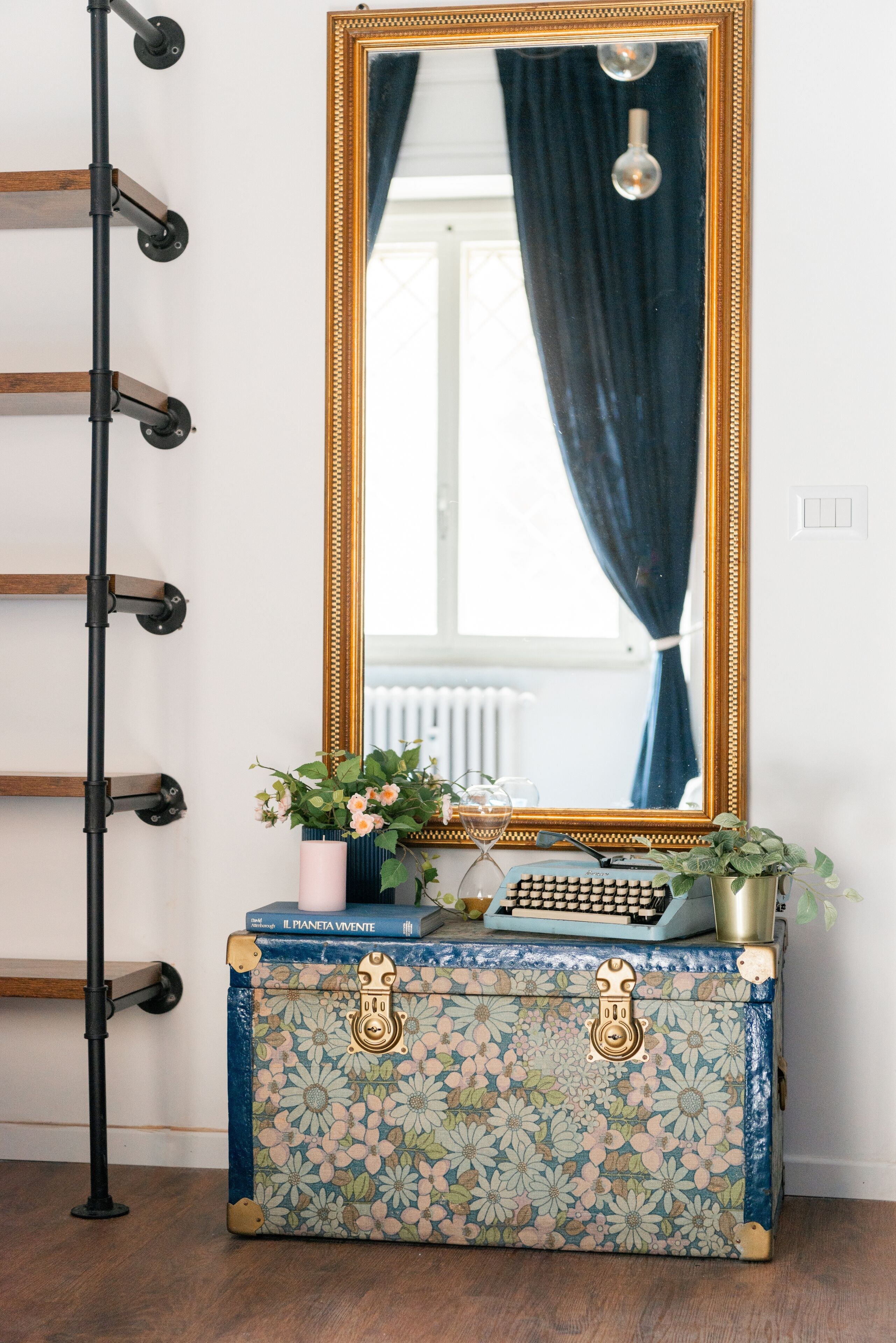 Una valigia vintage con motivi floreali e manici in ottone, posizionata su un pavimento di legno accanto a una rastrelliera in metallo nero.