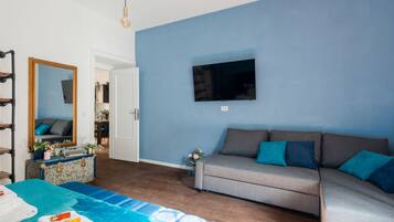 Ein Zimmer mit blauer Wand, einem grauen Sofa mit türkisfarbenen Kissen, einem flachbildfernseher, der darüber angebracht ist, einem Holzboden und einem großen Spiegel mit einer floralen Kommode.