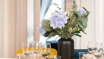 Ein Esstisch ist gedeckt mit Tellern, Gläsern und Besteck, eine Vase mit Blumen und ein blauer Stuhl.
