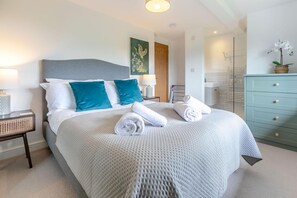 Een netjes opgemaakt bed met witte en teal kussens, een nachtkastje met een lamp, en een blauwe dressoir met een potorkidee.