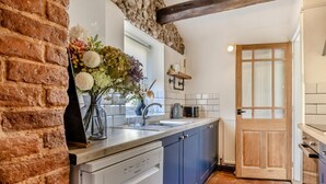 Une cuisine équipée d’un lave-vaisselle, d’un évier et d’une cuisinière. Un mur en briques se trouve d’un côté et une porte en bois de l’autre.