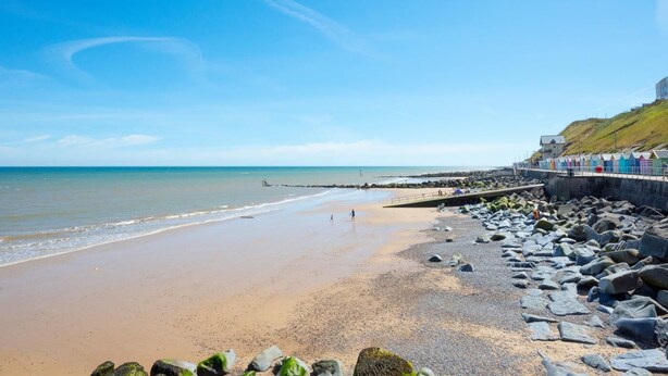 Une plage bordée d’une côte rocheuse, une promenade agrémentée de cabanes de plage aux couleurs vives et un ciel bleu dégagé.