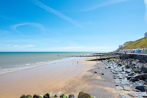 Ein Strand mit felsiger Küste, eine Promenade mit bunten Strandkörben und einem strahlend blauen Himmel.