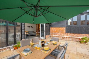 Un coin repas avec une table dressée pour quatre personnes, entourée de chaises, sous un parasol vert.