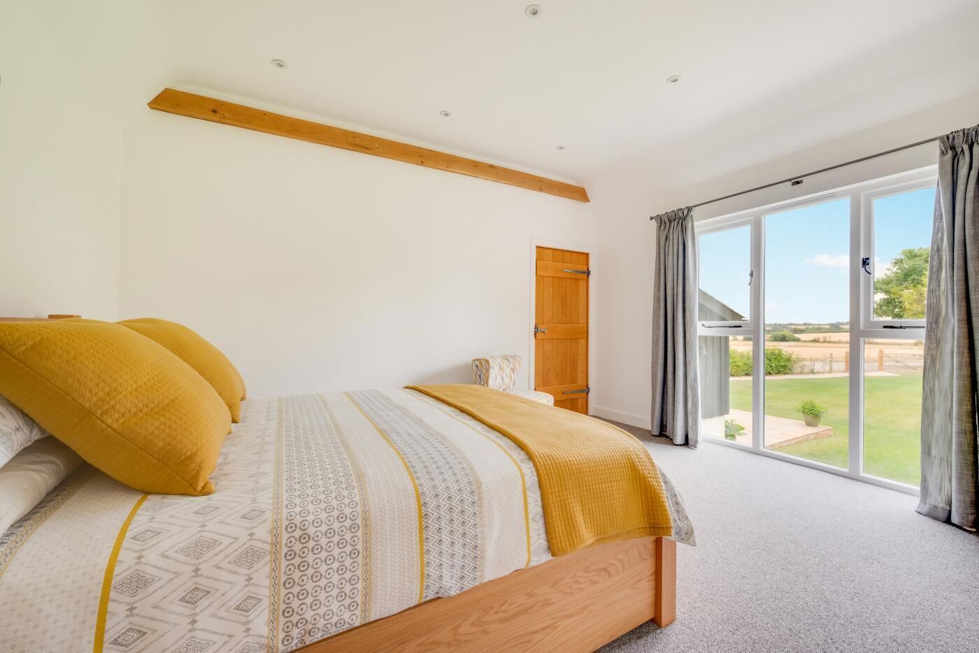 A bedroom with a large bed, a wooden ceiling beam, a sliding glass door to a garden, and a wooden door.