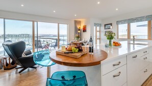 Een moderne keuken met een groot eiland, een eethoek met een glazen tafel en uitzicht op een jachthaven.