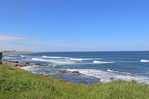 A coastal landscape with waves crashing onto rocks, a clear blue sky, and a distant building on the horizon. - 3 Bed in Seahouses (oc-s34113) (Seahouses)