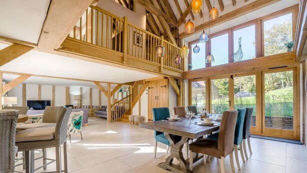 A spacious dining area with a wooden table and chairs, a staircase leading to an upper level, and large windows offering a view of the outdoors. - 2 Bed in Holmbury St. Mary (oc-midfb) (Holmbury Saint Mary)