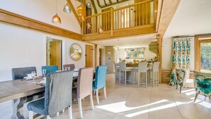 A spacious dining area with a high ceiling and wooden beams, a kitchen in the background, and a view of the outdoors through a window with curtains. - 2 Bed in Holmbury St. Mary (oc-midfb) (Holmbury Saint Mary)