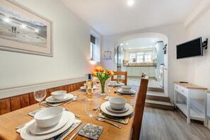 A dining area with a wooden table set for four, white plates, glasses, and a vase of tulips. A kitchen is visible in the background with a washing machine. - 4 Bed in Saundersfoot (oc-pw9275) (Saundersfoot)