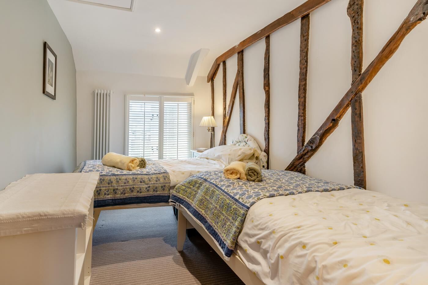 Une chambre avec deux lits, une fenêtre avec des stores, un radiateur et une structure en poutres apparentes.