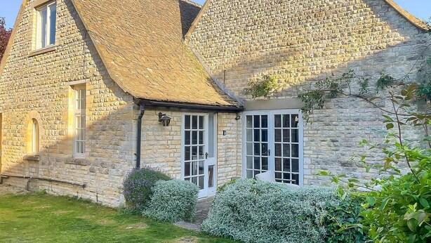 Un bâtiment en pierre avec un toit de chaume, une porte blanche et un petit jardin avec de l’herbe verte et des arbustes.