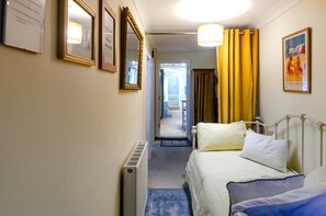 Habitación de hotel con cama, radiador, puerta que da a otra habitación y pasillo con espejo y diplomas enmarcados.