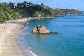A coastal beach with a rocky outcrop, clear blue water, and houses on a hillside. - 4 Bed in Tenby (oc-pw9281) (Tenby)