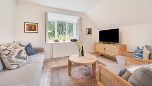 Ein modernes Wohnzimmer mit einer Couch, einem Couchtisch mit Vase und Gläsern, einem Fernseher auf einer Holzkommode und einem Fenster mit Blick auf Grünflächen.