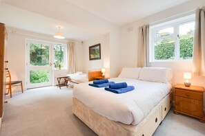 Une chambre d’hôtel avec un grand lit, un bureau et une chaise. Il y a des fenêtres avec des rideaux, un tableau au mur et une vue sur la verdure à l’extérieur.
