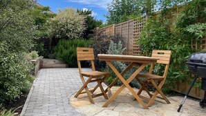 Eine Terrasse mit zwei Holzstühlen und einem Tisch, umgeben von einem Garten mit verschiedenen Pflanzen und einem Holzzaun.