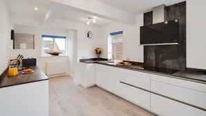 A modern kitchen with white cabinets, a black countertop, and a large black stove. There is a window with blue blinds and a small sailboat decoration on the windowsill. - 4 Bed in Sandsend (oc-wa330) (Sandsend)