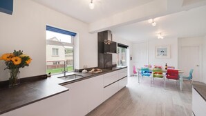 A modern kitchen with a large island, a dining area with colorful chairs, and a view of a house through a window. - 4 Bed in Sandsend (oc-wa330) (Sandsend)