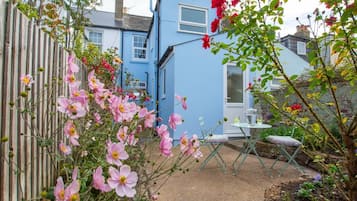 A garden with pink flowers, a blue house, and a small table with chairs.