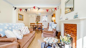 Un salon chaleureux avec un canapé, un fauteuil et une petite table. On y trouve une cheminée, un coin repas avec des chaises et un escalier menant à un autre niveau.