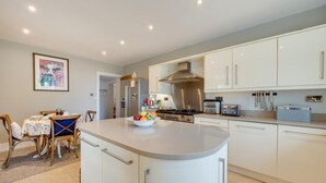 A modern kitchen with white cabinets, a central island with a bowl of fruit, a stove, and a dining area with a table and chairs. - 4 Bed in Brithdir (oc-wag722) (Brithdir)