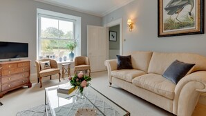 A living room with a beige sofa, a glass coffee table, a wooden TV stand, and a large window. - 4 Bed in Brithdir (oc-wag722) (Brithdir)