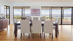 Uma sala de jantar com mesa e cadeiras, janelas amplas com vista e um quadro na parede.