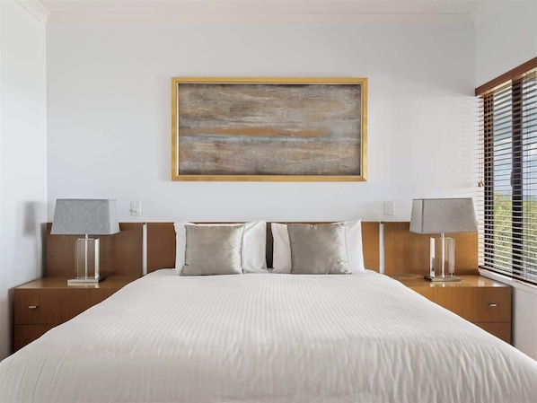A hotel room with a large bed, two bedside tables, a painting on the wall, and a window with blinds.