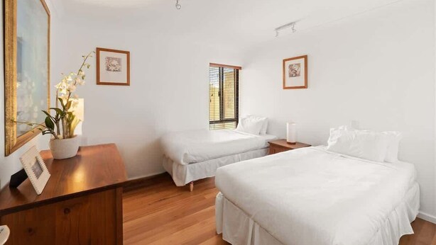 Quarto de hotel com duas camas de solteiro, uma escrivaninha de madeira, um vaso com planta e duas fotos emolduradas nas paredes.