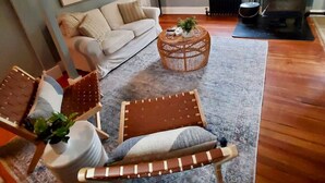 Een woonkamer met een bank, een rond rieten bijzettafeltje, een houten stoel en een open haard.