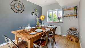 Un coin repas avec une table en bois dressée pour six personnes, une horloge moderne accrochée au mur et une fenêtre donnant sur un paysage verdoyant.
