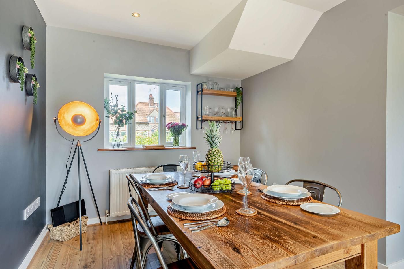 A dining area with a wooden table set for four, a large window with a view, and a floor lamp.