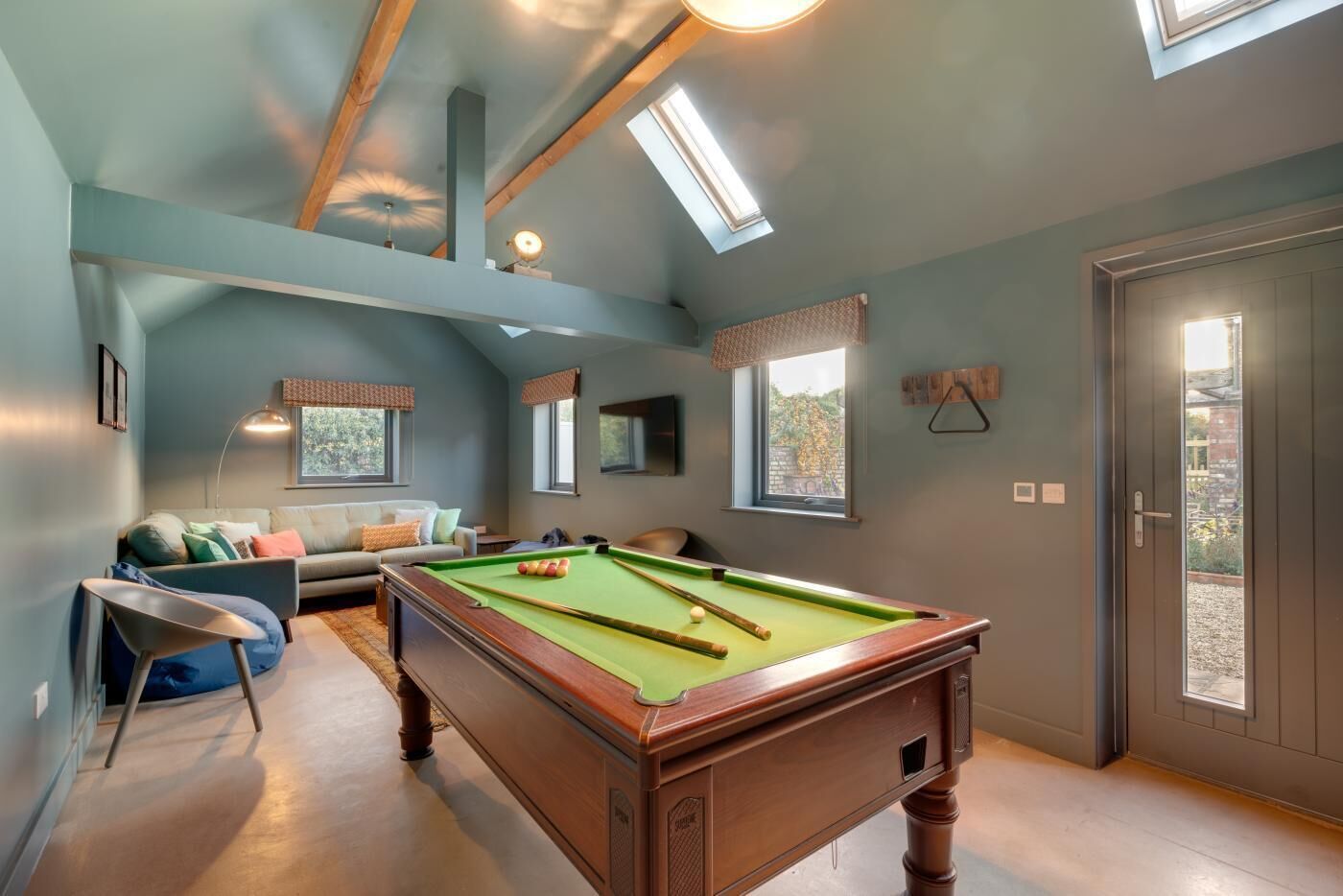 Une table de billard dans une pièce avec un plafond haut, un canapé et une porte donnant sur l’extérieur.