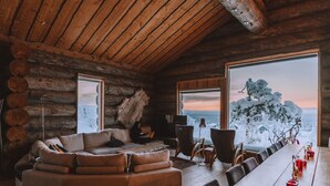 Interior de una cabaña de troncos con un sofá grande, una mesa de comedor con sillas y vistas a un paisaje nevado a través de ventanas amplias.