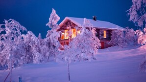 Una cabaña de troncos con techo nevado y ventanas, rodeada de árboles cubiertos de nieve.